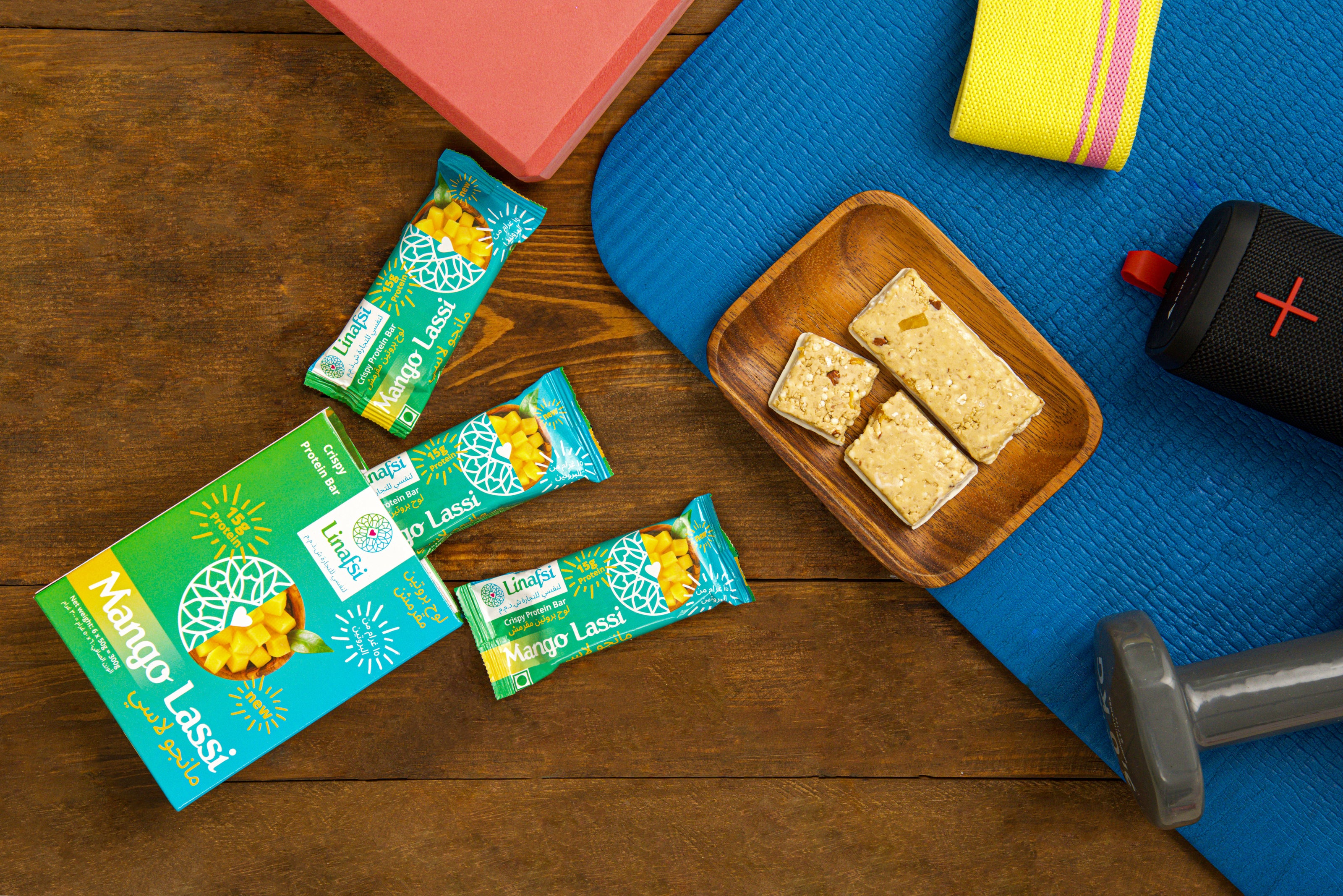 Linafsi Mango Lassi protein bars staged in a fitness setting with a blue yoga mat, dumbbell and exercise band. 