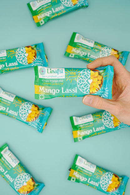 A hand holding a mango lassi protein bar with other bars in the background on a blue surface. 